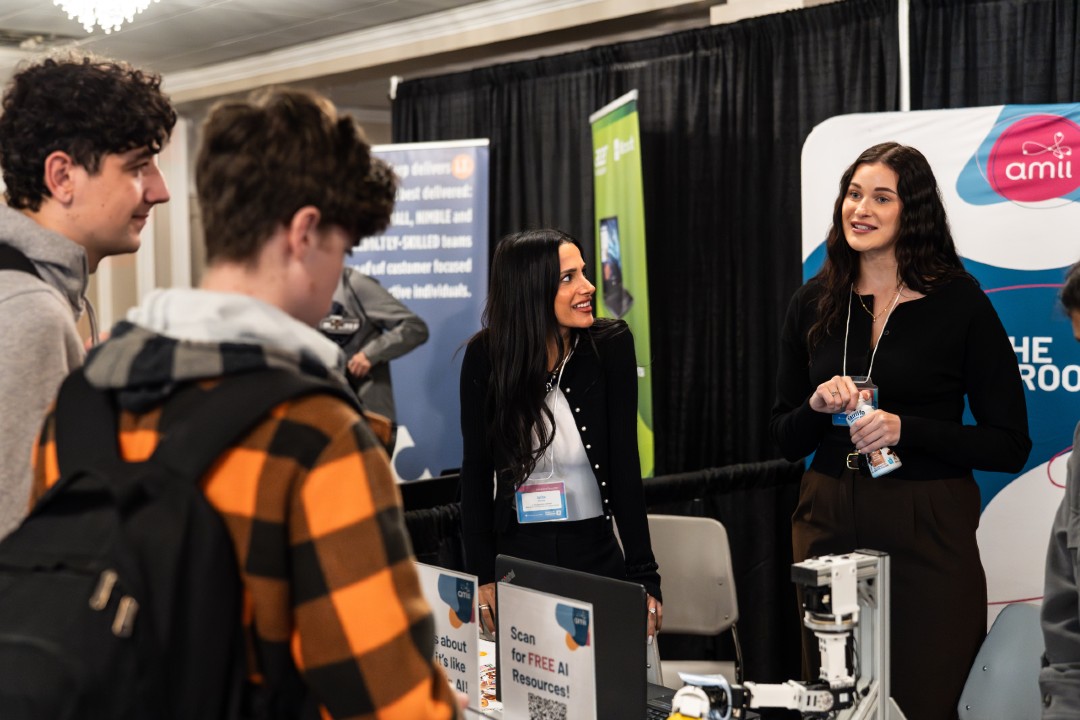 Upper Bound focuses on AI literacy to better prepare future workers Two adults at a booth with an Amii banner talk to two high school students.