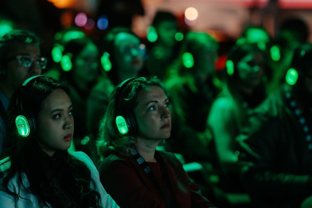 A seated conference audience wearing headphones that glow with green light.
