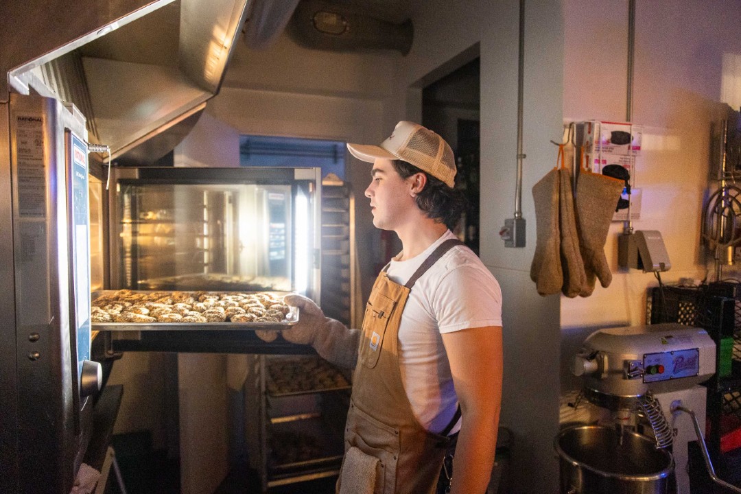 Meuwly's drops retail to cook up new food brands A baker wearing a trucker hat and a Carhartt apron removes a tray of bagels from an industrial oven.