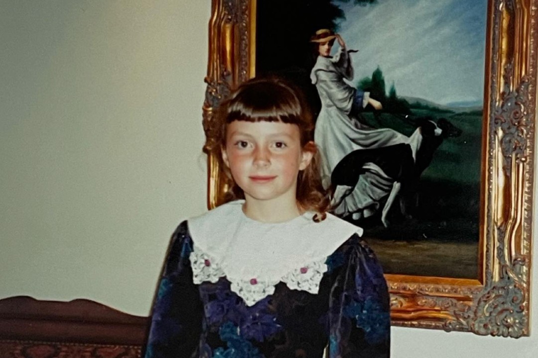 A young girl poses for a photo in front of a painting of someone walking two dogs.