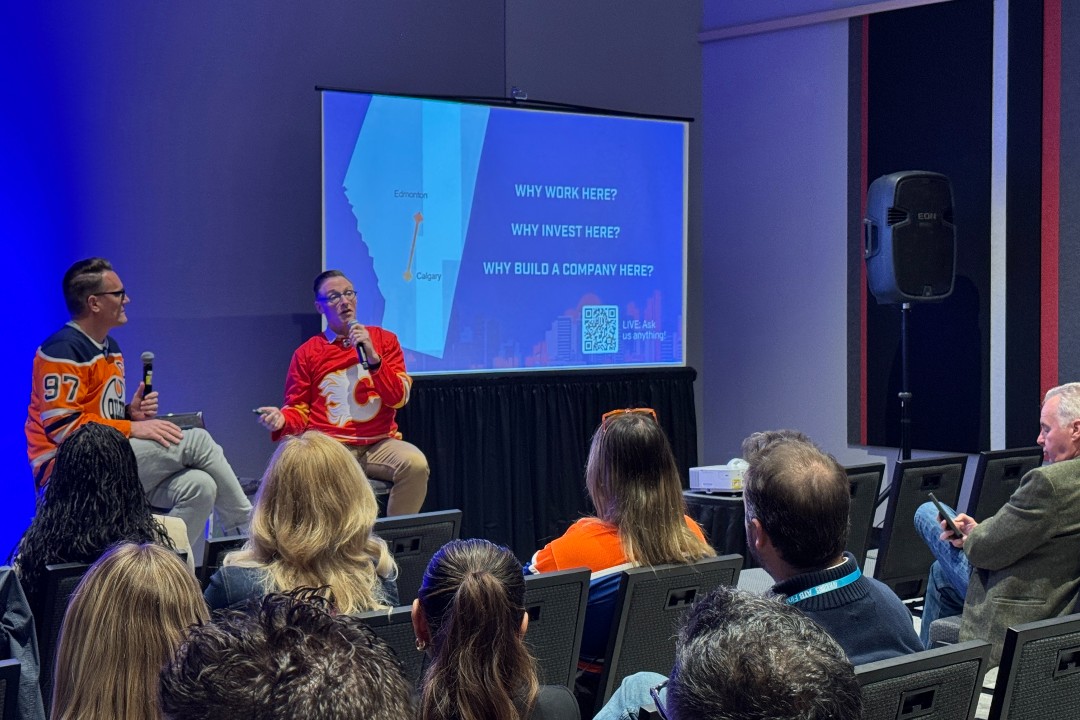 Edmonton forms new partnership with Calgary to strengthen tech in both cities Two seated men wearing hockey jerseys address an audience next to a screen that displays a map of Alberta with text that asks "Why work here?" among other questions.