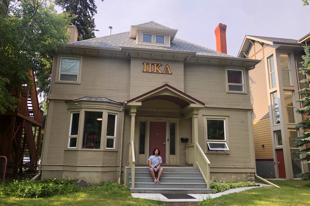 A man sits on the steps of a large beige two-storey house with Greek letters over the door.