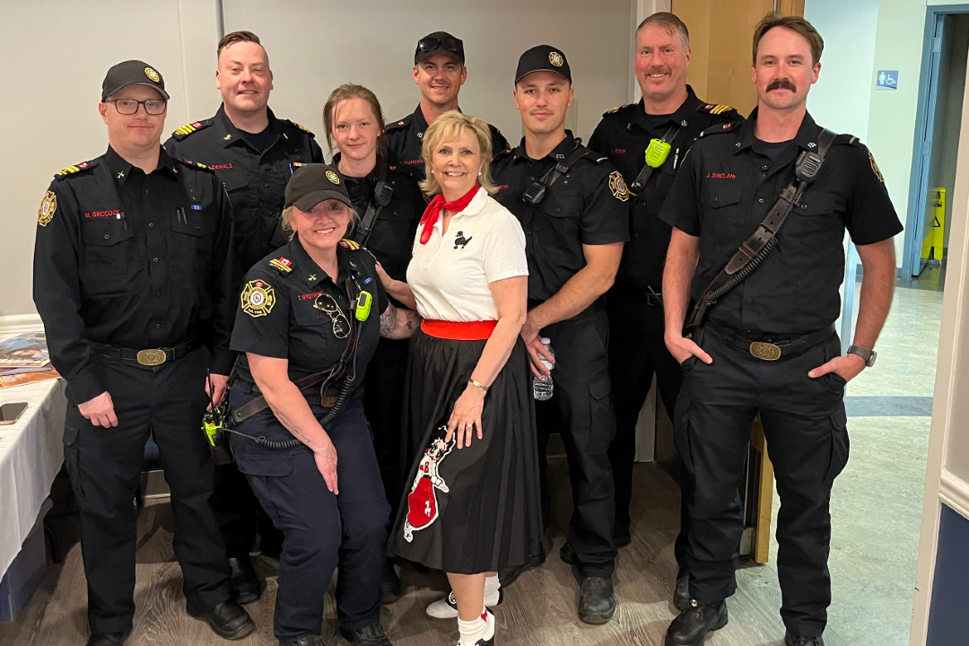 A group of eight firefighters pose for a photo with a woman wearing red, black, and white at the centre.]