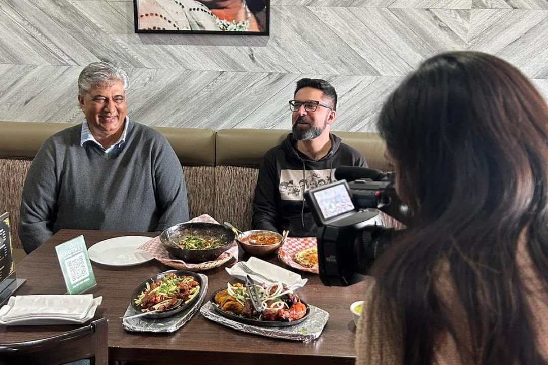 Diners sit at a table filled with South Indian food as a camera person looks on.