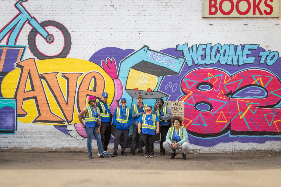 A group of seven people wearing blue-and-yellow safety vests pose in front of a colour mural in Old Strathcona.