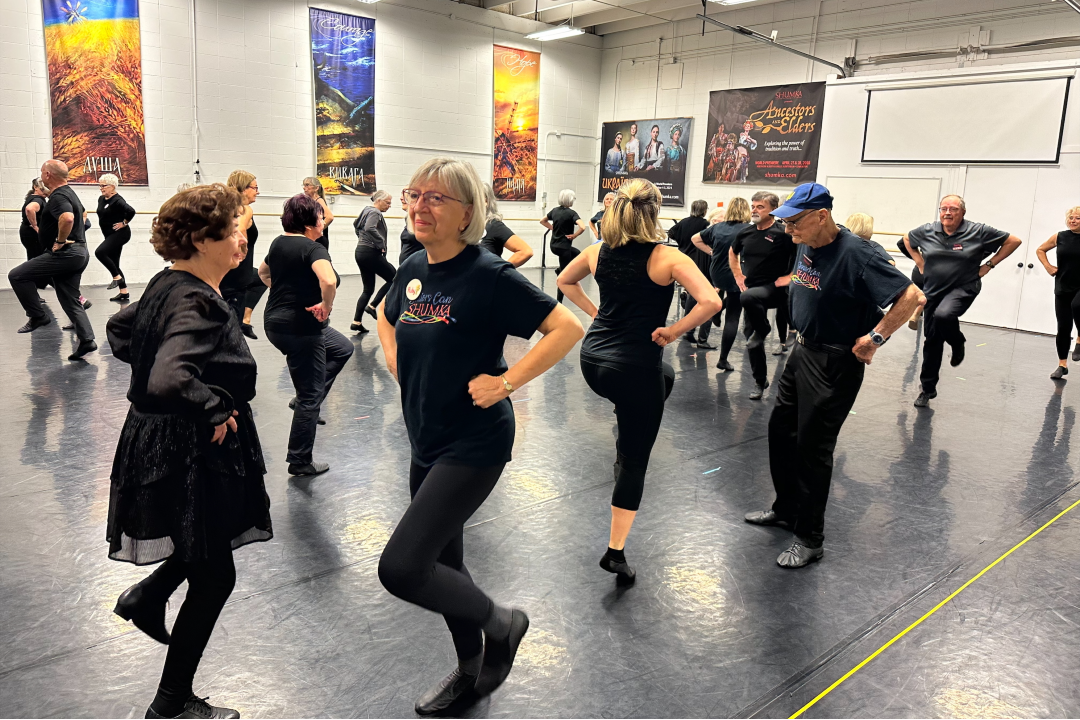 Dozens of dancers in black perform a partner dance in a studio.