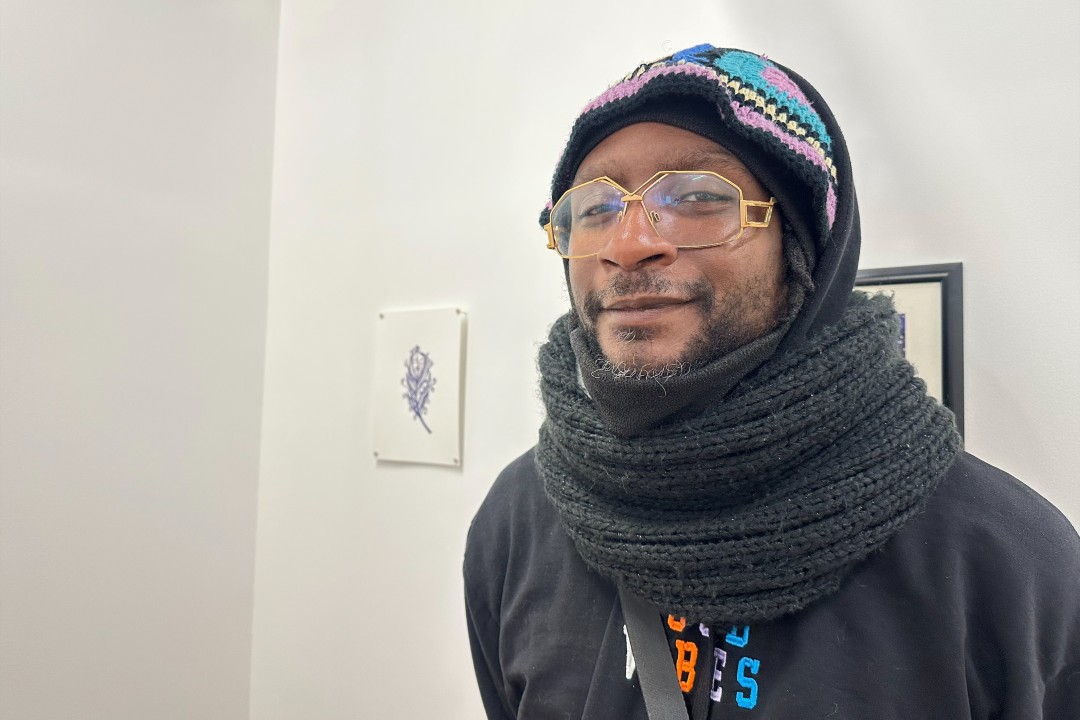 A man wearing a hat and scarf stands in front of art pieces.