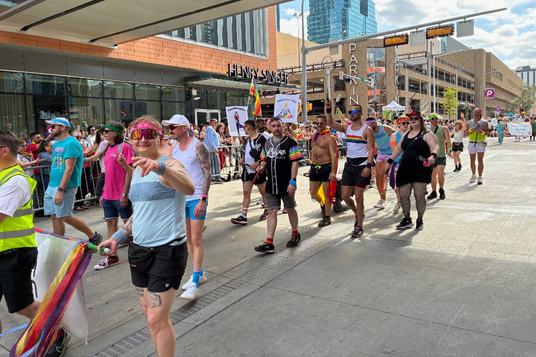 About two dozen people march, many waving rainbow flags, with spectators lining the street.