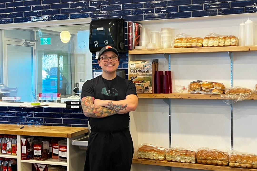 A smiling man crosses his arms next to hot dog and hamburger buns inside a meat shop.