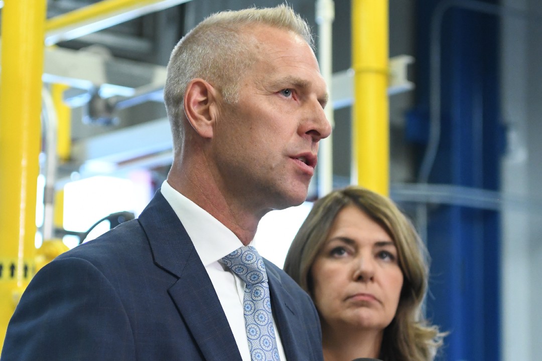 A man in a suit speaks at a podium while a woman looks on.