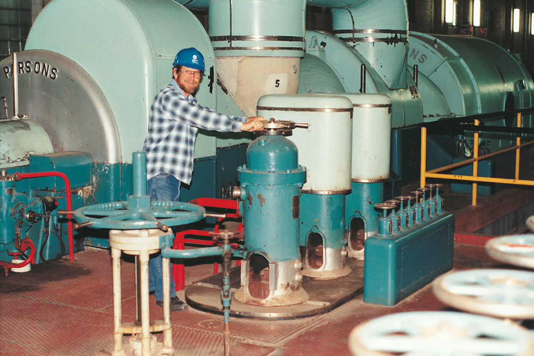 A smiling, bearded man wearing a hard hat grasps the top of a piece of equipment in front of a set of turbines