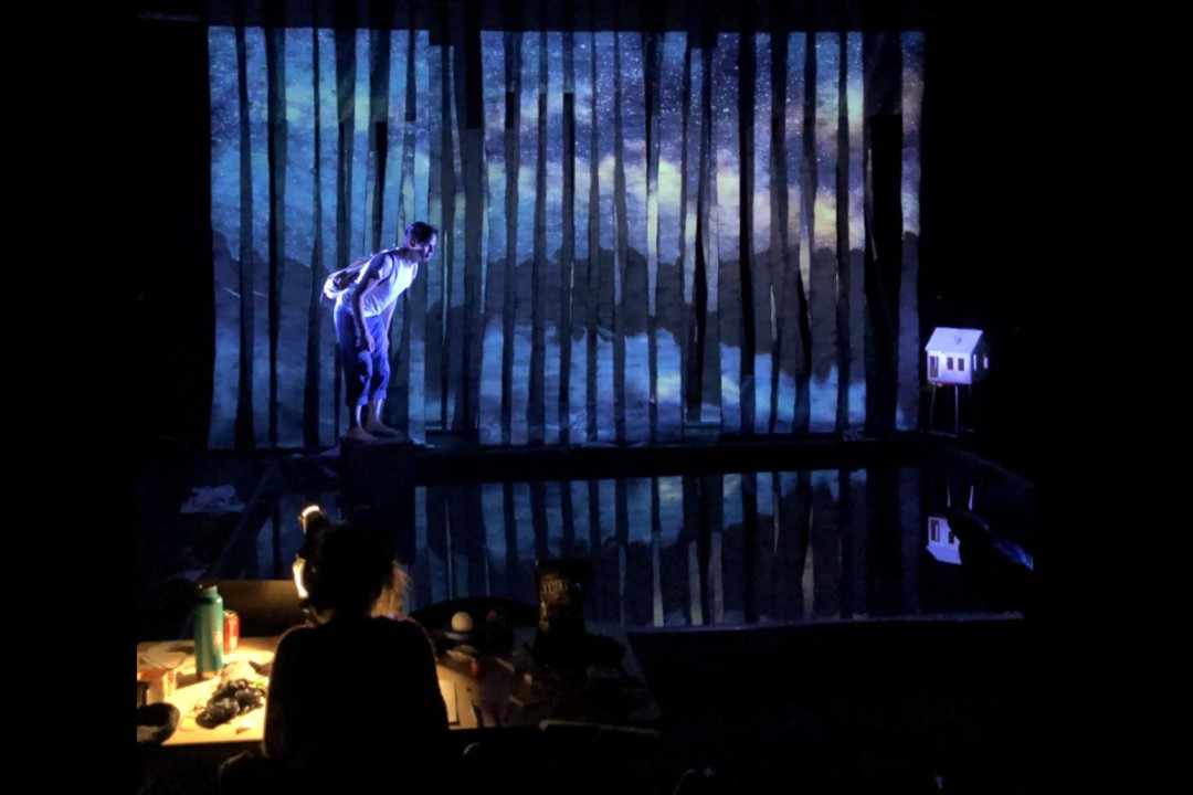 An actor stands on a stage while a stage manager looks on.