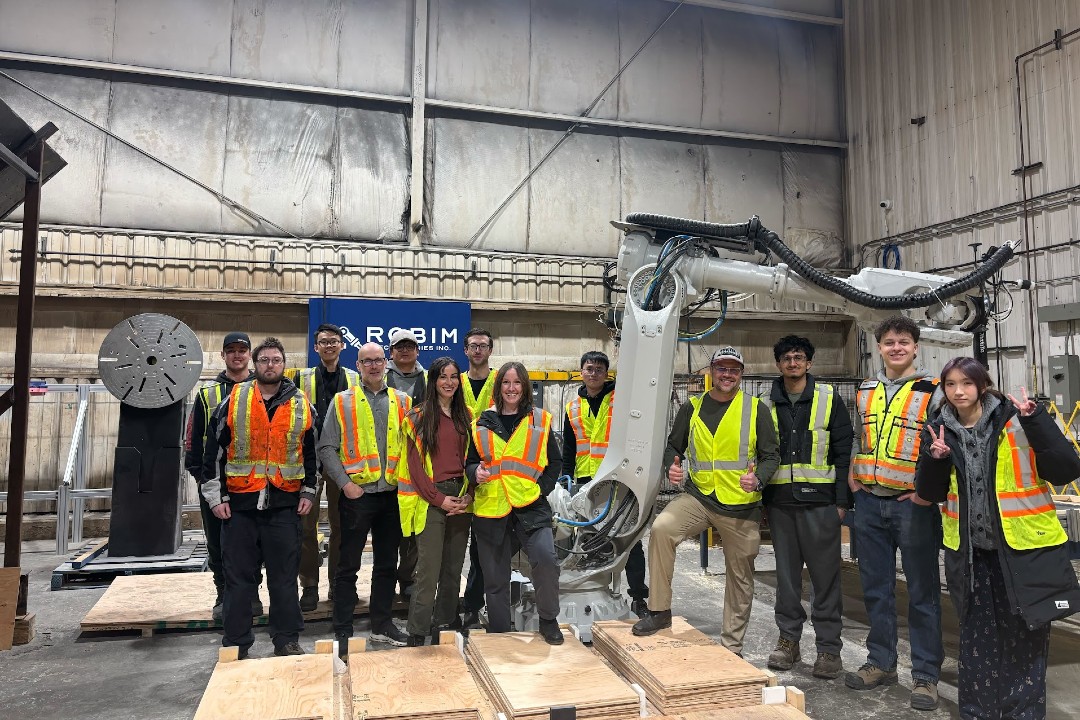 A group of people pose in safety vests pose near a robotic arm inside a warehouse.
