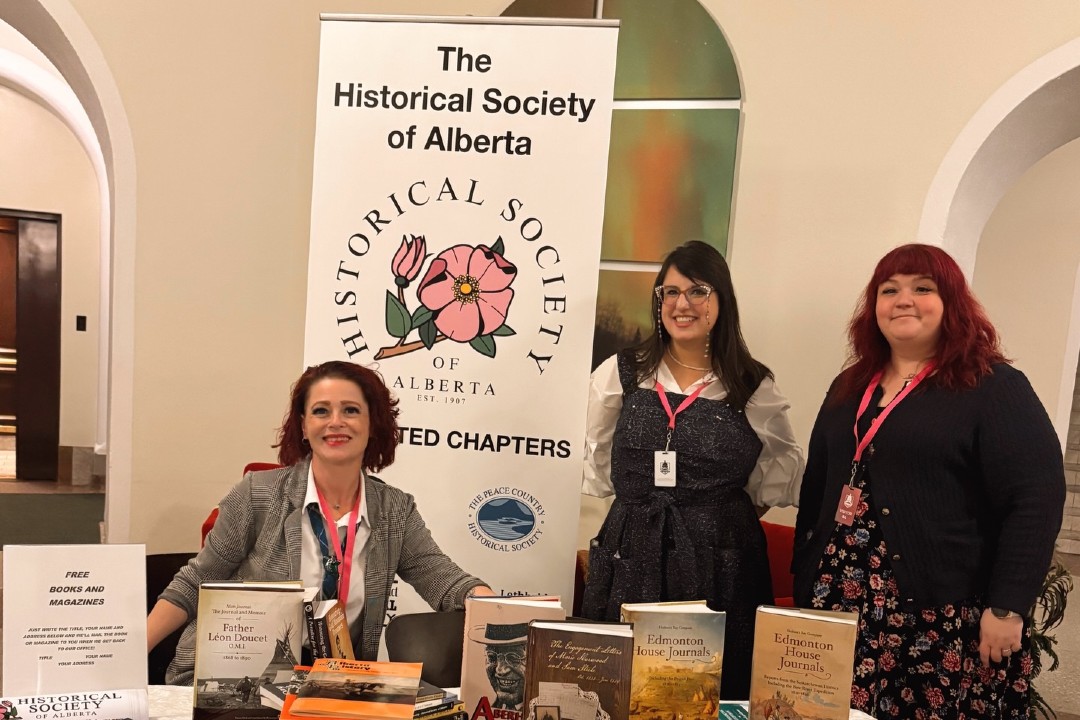 Three people gathered around a table covered in books and a banner that reads The Historical Society of Alberta