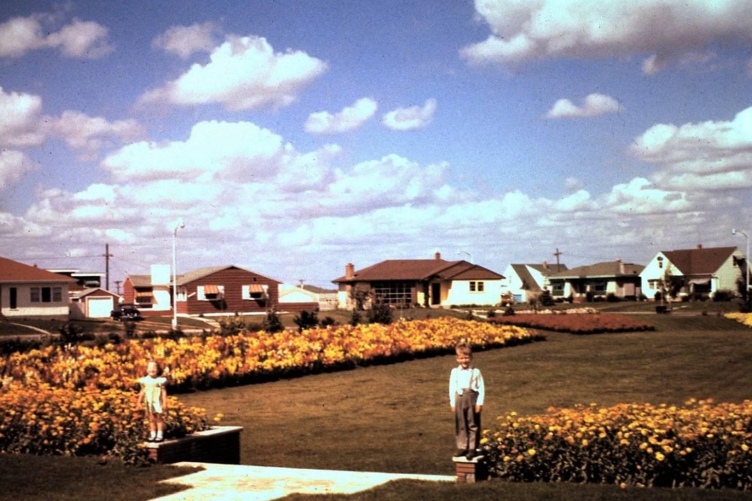 Two children stand on a wall in a park. In the background are bungalows and yellow flowers.