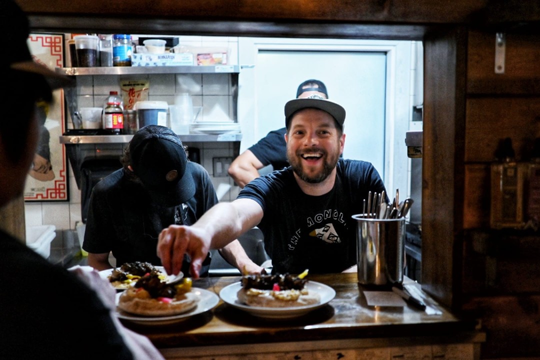 A smiling man passes food dishes from a restaurant kitchen to waiting servers.