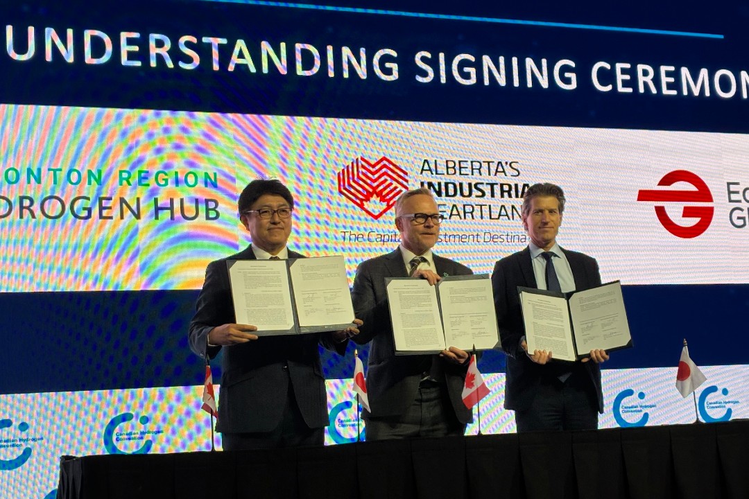 Three men in suits hold up a signed memorandum of understanding on a stage.