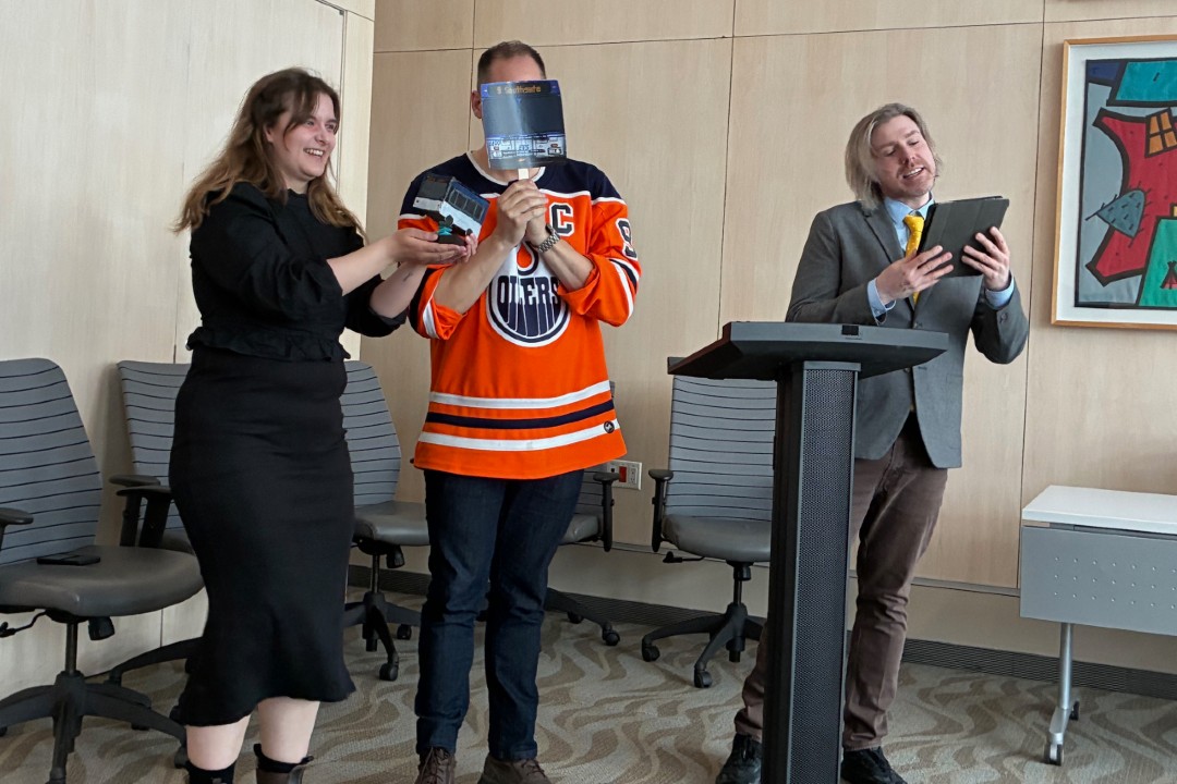 Three people stand behind a podium, one holding a bus mask in front of his face.