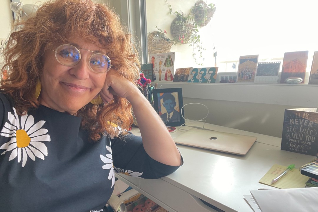 A smiling woman sits at her desk, resting her head on her hand