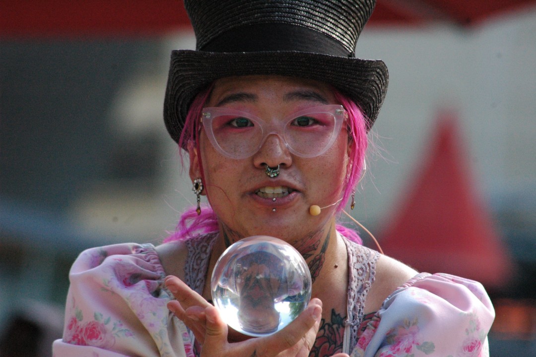 A woman in a top hat holds a small crystal ball, reflecting an image of the crowd upside down