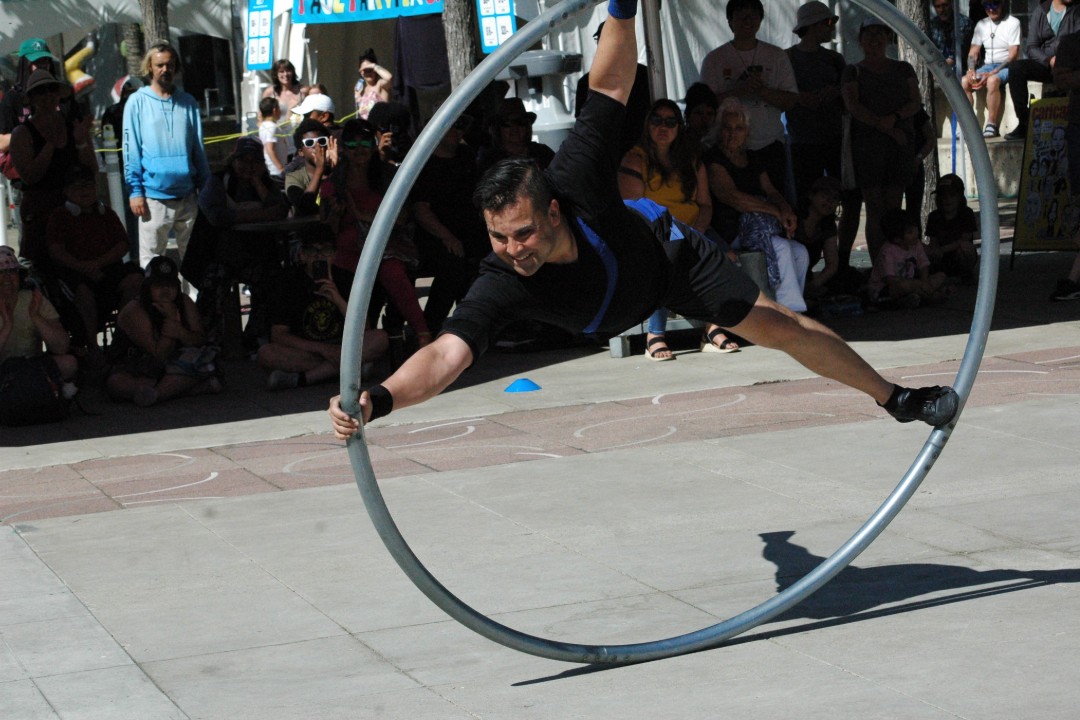 A smiling man balances in a large metal ring while spectators watch