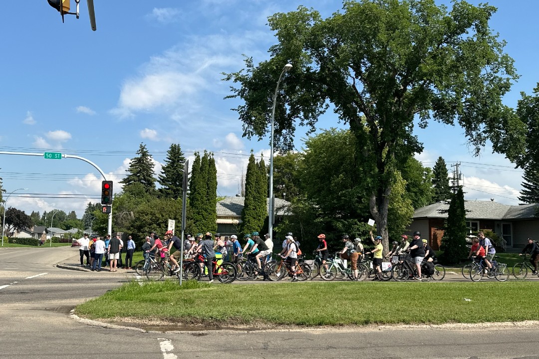 About 60 cyclists ride past a group of people on the sidewalk