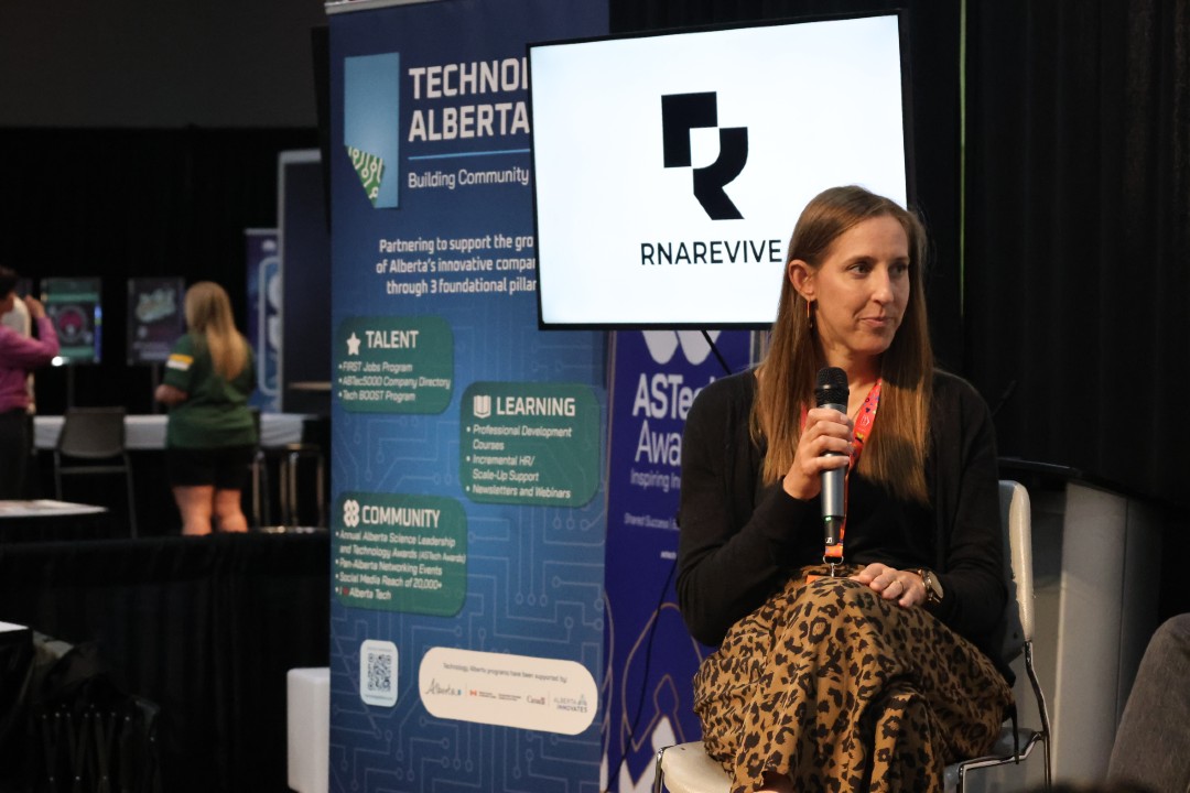 A woman seated on a stage with backdrops that read RNARevive and Technology Alberta