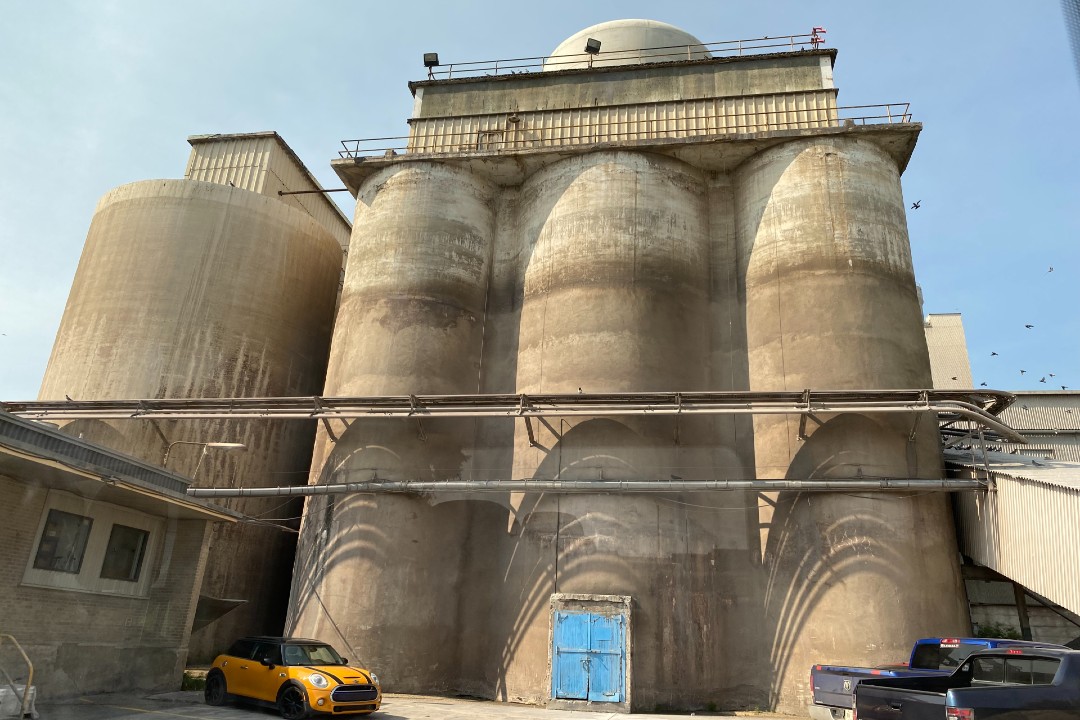 Four concrete cylinders loom over a yellow Mini Cooper and two blue trucks. A closed blue door resides in the centre of one of the cylinders. Dirt and grime sticks to the outer walls of the cement cylinders.