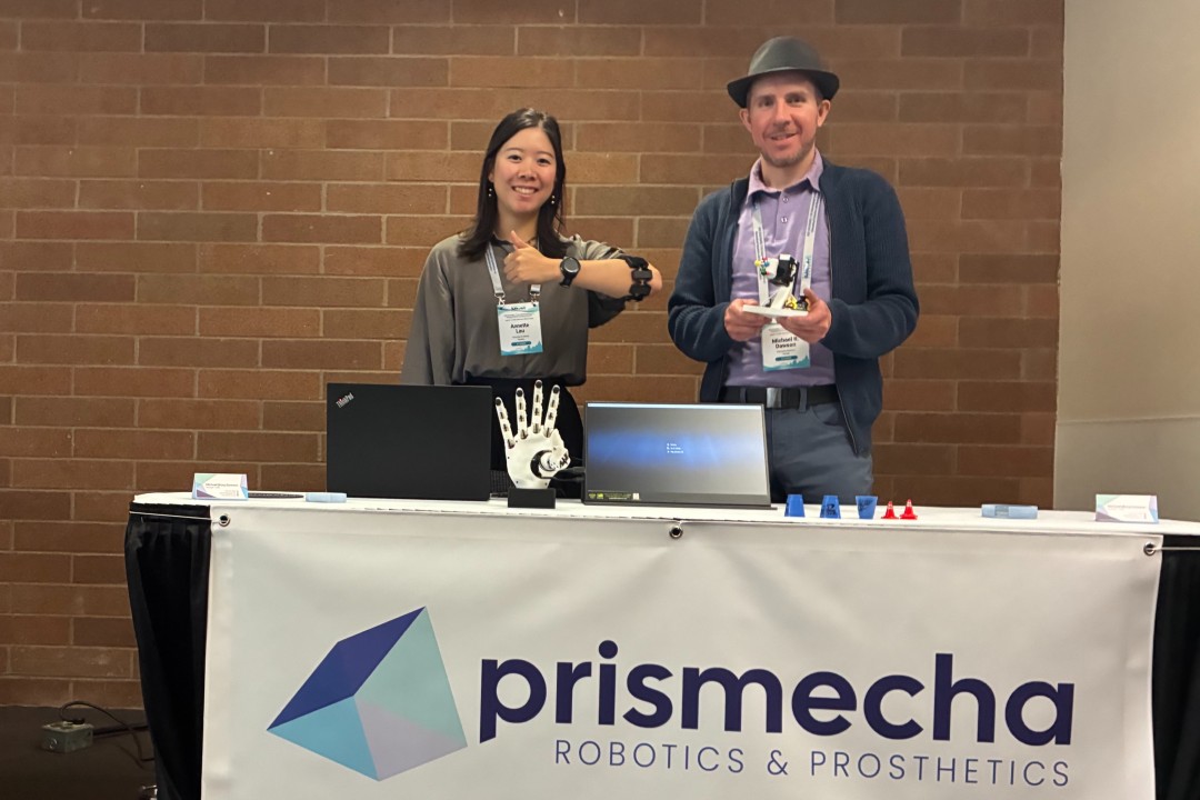 Two people pose with robots behind a booth that says Prismecha Robotics &amp; Prosthetics