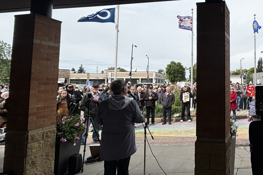 A speaker addresses a crowd of people in front of Sturgeon County Centre. Some members of the crowd are holding signs with anti-data centre messages.