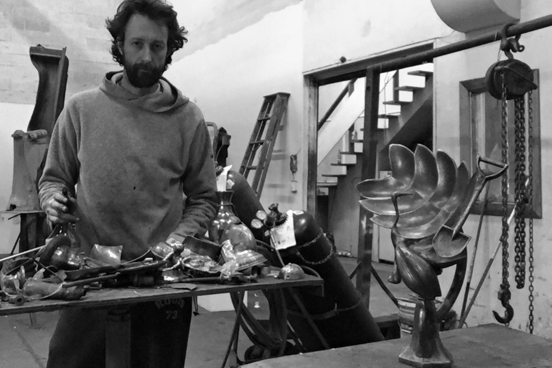 A black-and-white photograph of Ryan McCourt in a metalworking studio. Metal sculptures rest on a table in the foreground.