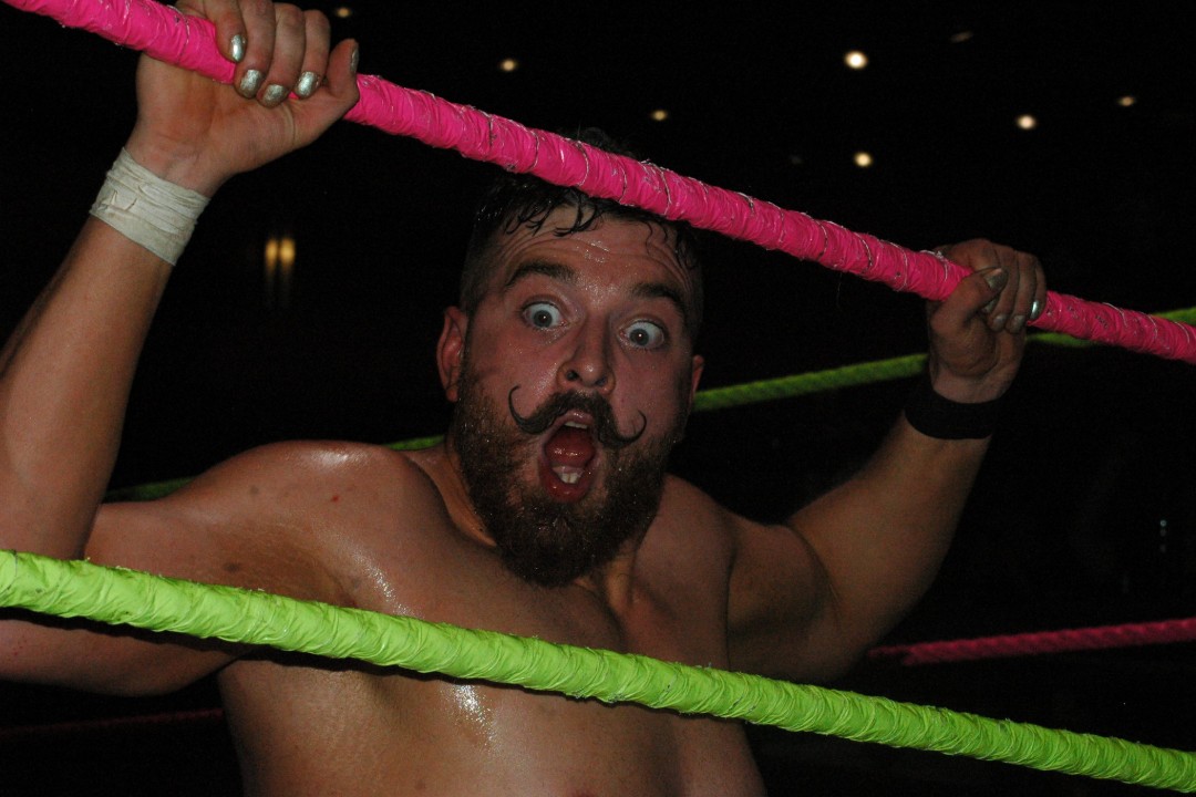 A moustachioed wrestler gestures while holding onto the ropes