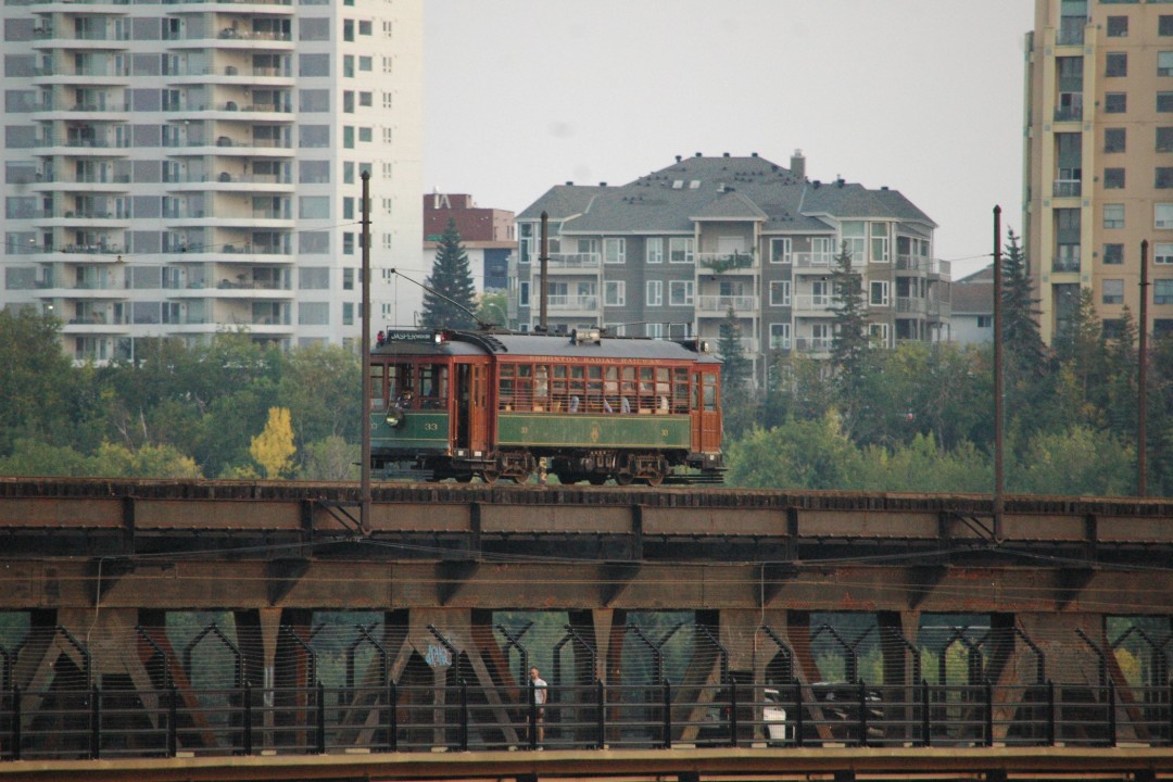 A streetcar travels across an iron bridge with trees and highrises in the background