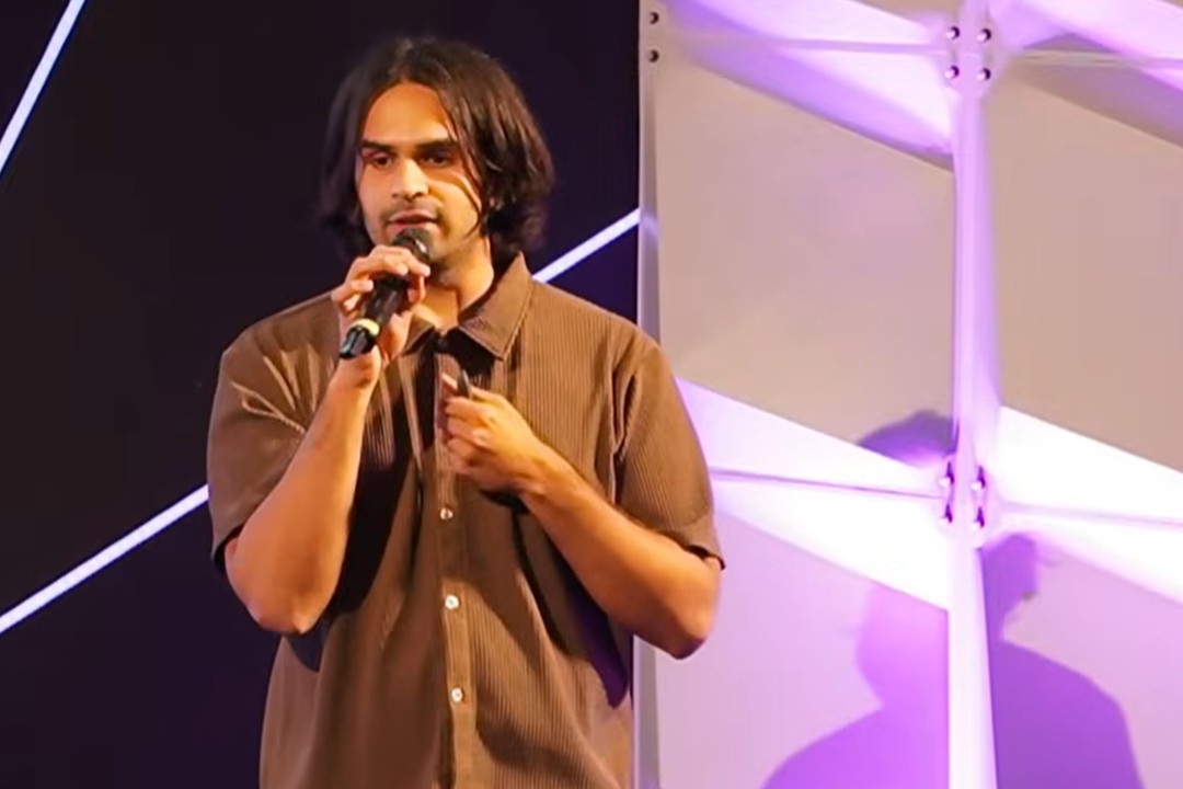 A man speaks into a microphone on a purple-lit stage