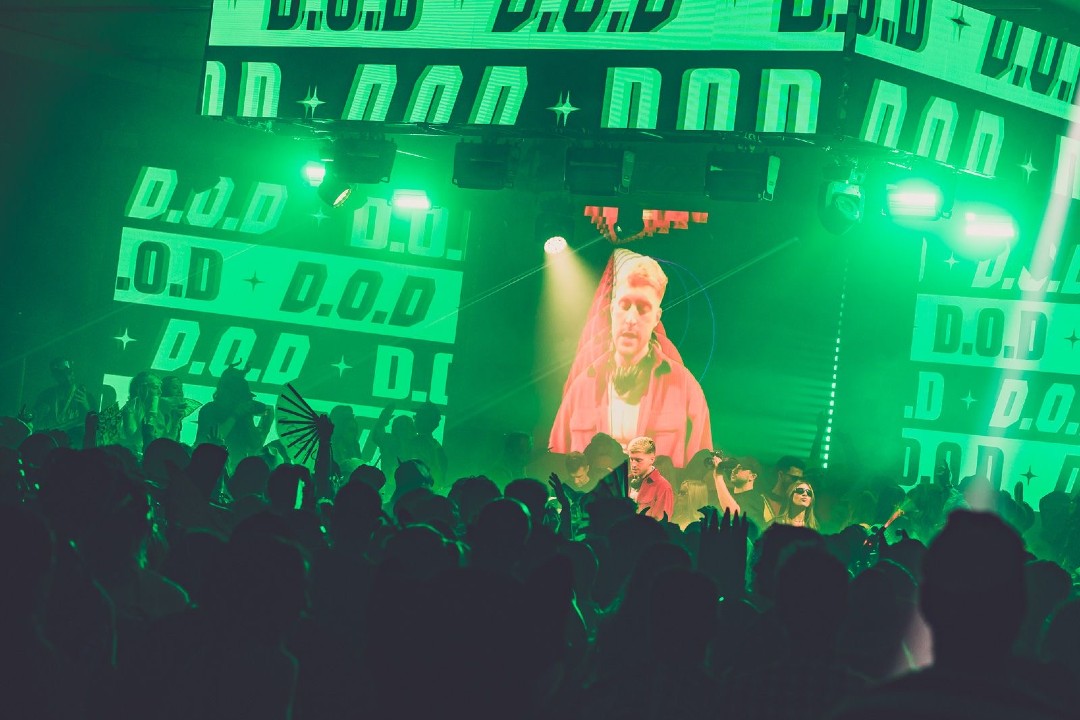 A DJ performs for an audience with green lighting and his name, D.O.D., displayed throughout the backdrop