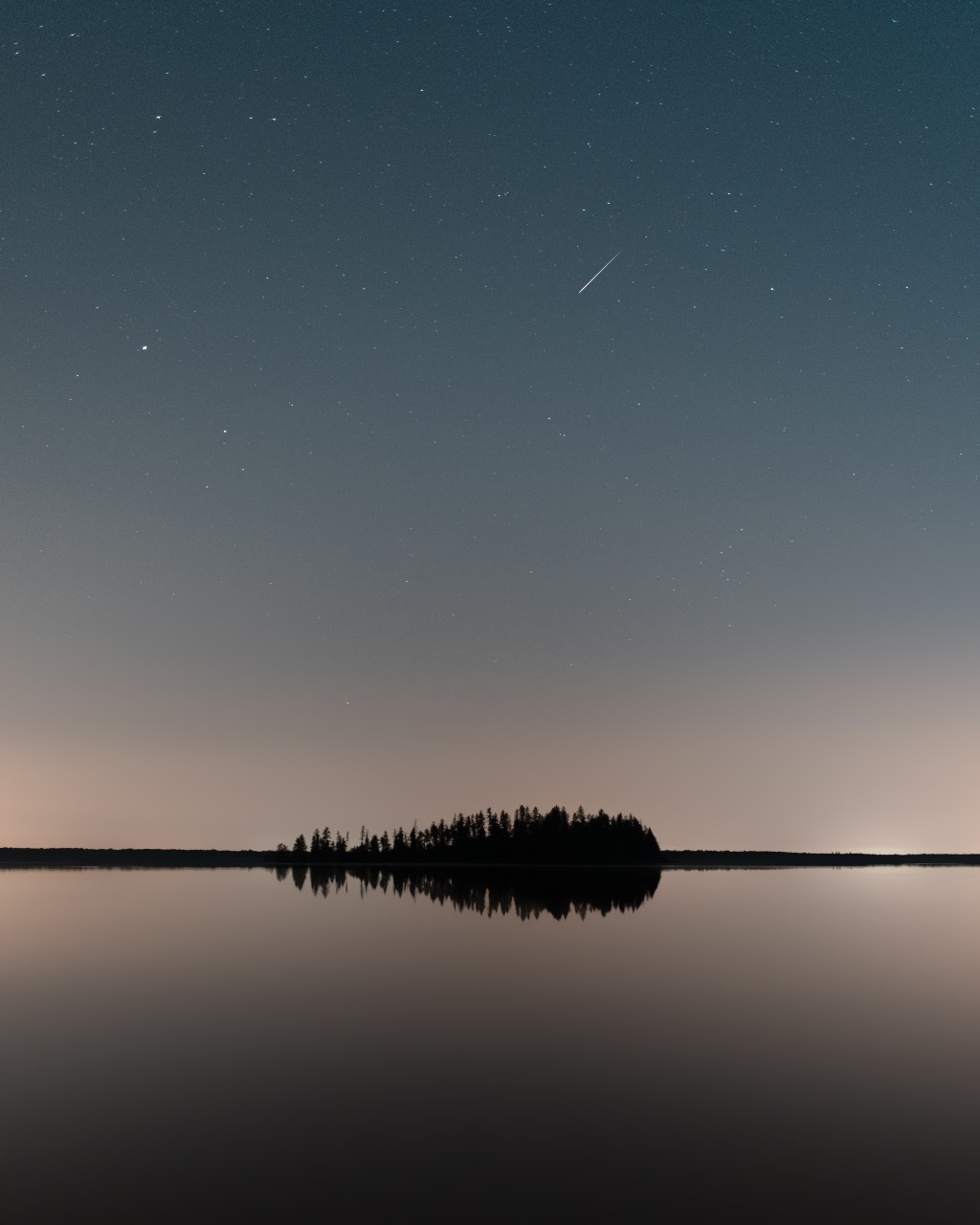 A star streaks through the twilight sky above a clump of trees in the middle of a body of water