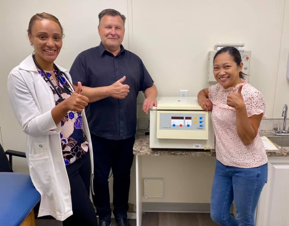 Three people in a medical clinic give a thumbs-up sign beside a centrifuge