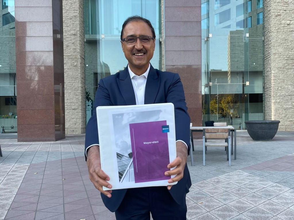 A smiling Amarjeet Sohi holds a white binder with the words "Mayor-Elect" on the front at City Hall